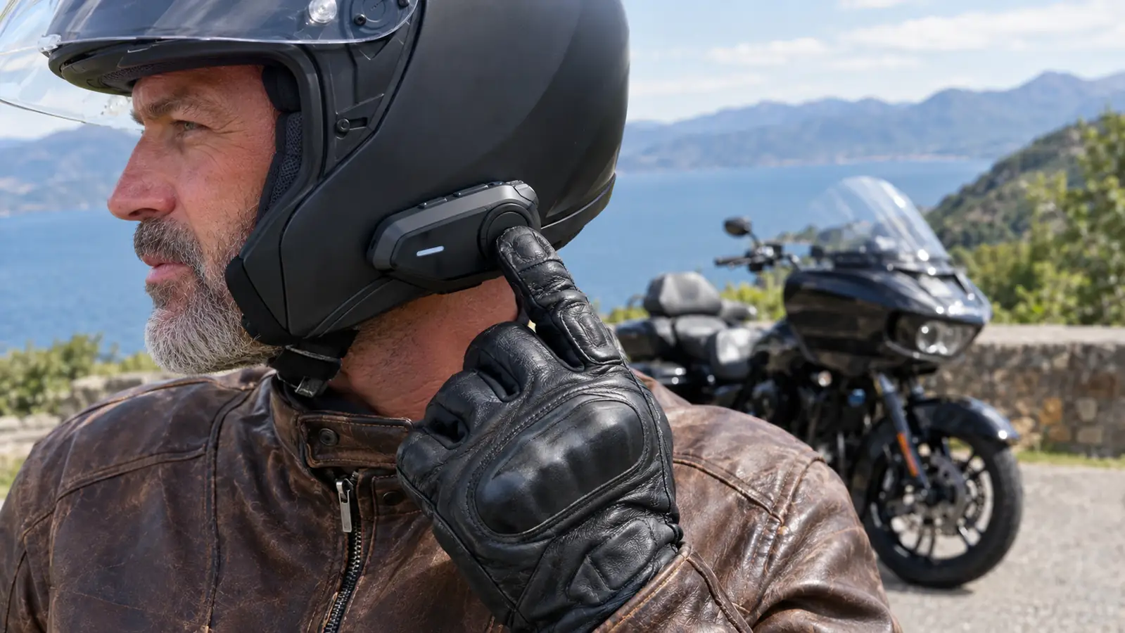 A rider's gloved hand pressing the main button on a helmet-mounted communicator beside a parked touring motorcycle