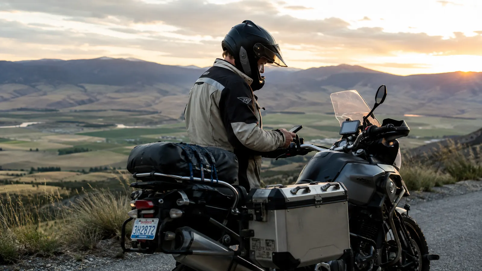A rider in a riding jacket and helmet checking a phone mounted on the handlebar, motorcycle parked at a scenic overlook