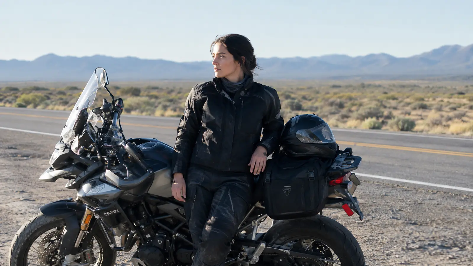Rider leaning against a touring motorcycle at a roadside stop, helmet resting on the tail bag