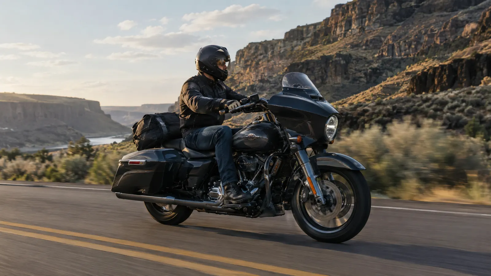 A touring motorcycle with a tail bag mounted behind the seat, riding a two-lane canyon road in late afternoon light.